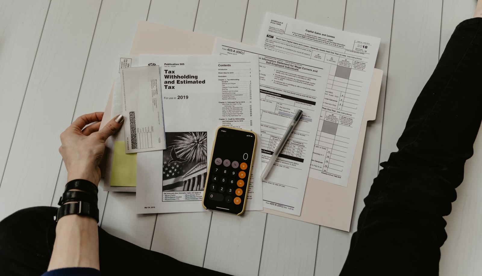 Desk spread with printed compliance forms, financial statements, a calculator and a smartphone showing populated rows of figures